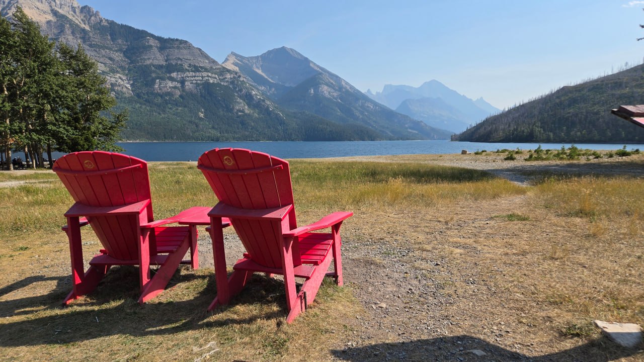 Two red chairs facing a lake framed by mountain peaks